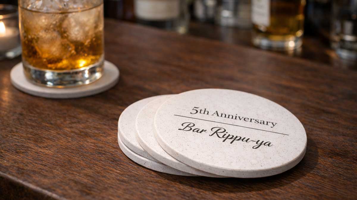 ◇代替テキスト 周年記念の粗品として配布される名入れコースターと小さなギフトが並ぶバーカウンターのイメージ ◇タイトル 周年記念 粗品|名入れコースターと記念品のイメージ ◇説明 飲食店の周年記念でお客様に配布する粗品をイメージした写真。名入れされた珪藻土コースターと小さなギフトがバーカウンターに並び、感謝の気持ちを伝える実用的な記念品の雰囲気を表現している。