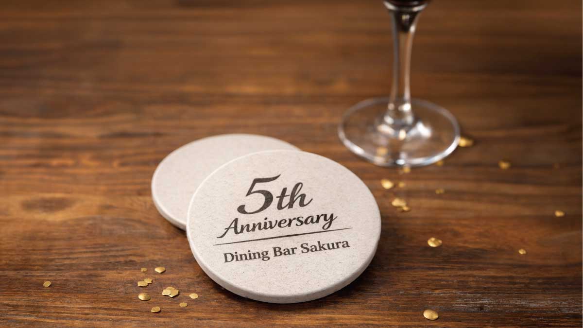 店名と周年が刻まれた薄型の珪藻土コースターが2枚重なって置かれた記念品イメージ